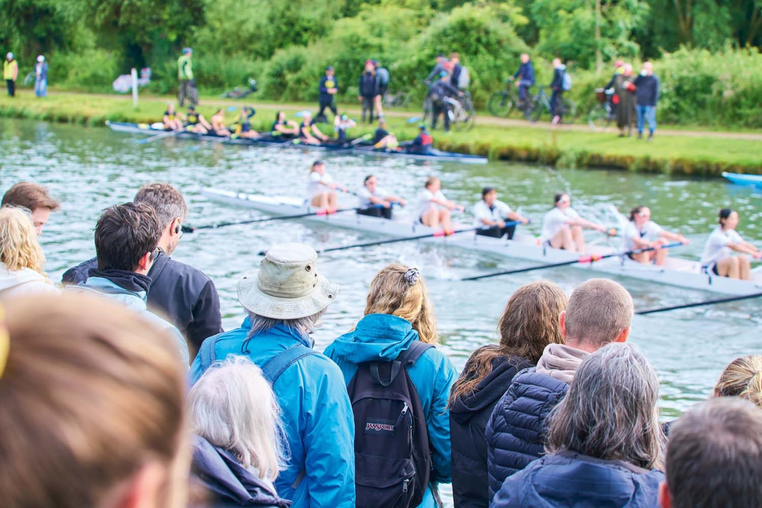 Students in boat on river out of focus. Crowd in focus on bank.