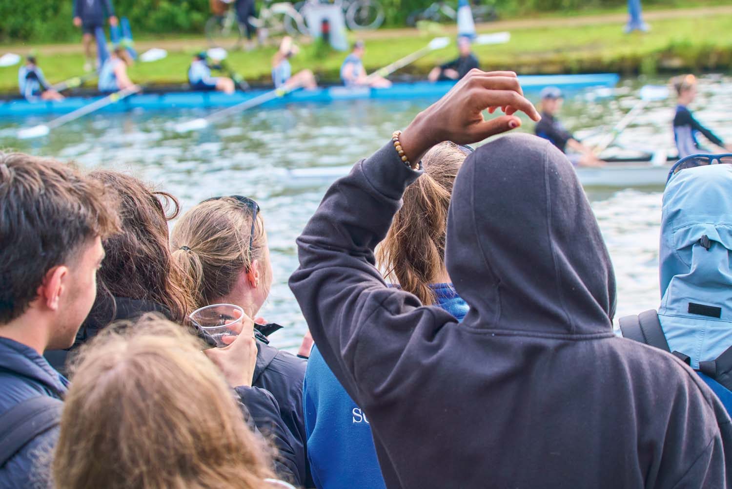 Students in boat on river out of focus. Crowd in focus on bank.