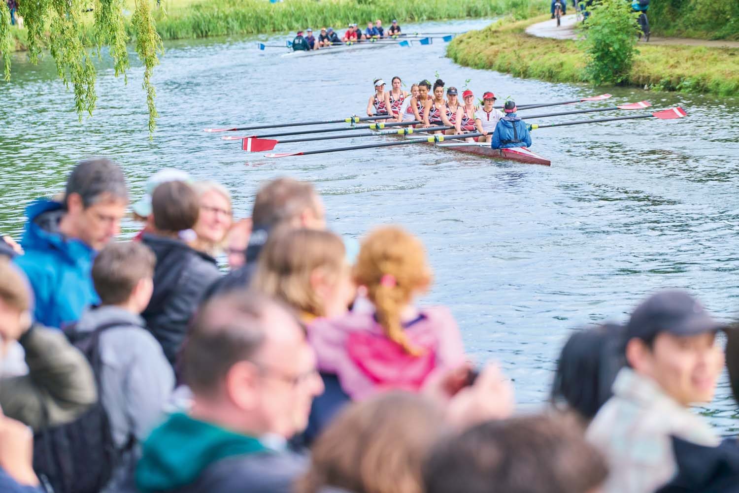 Students in boat on river. Crowd out of focus on bank.